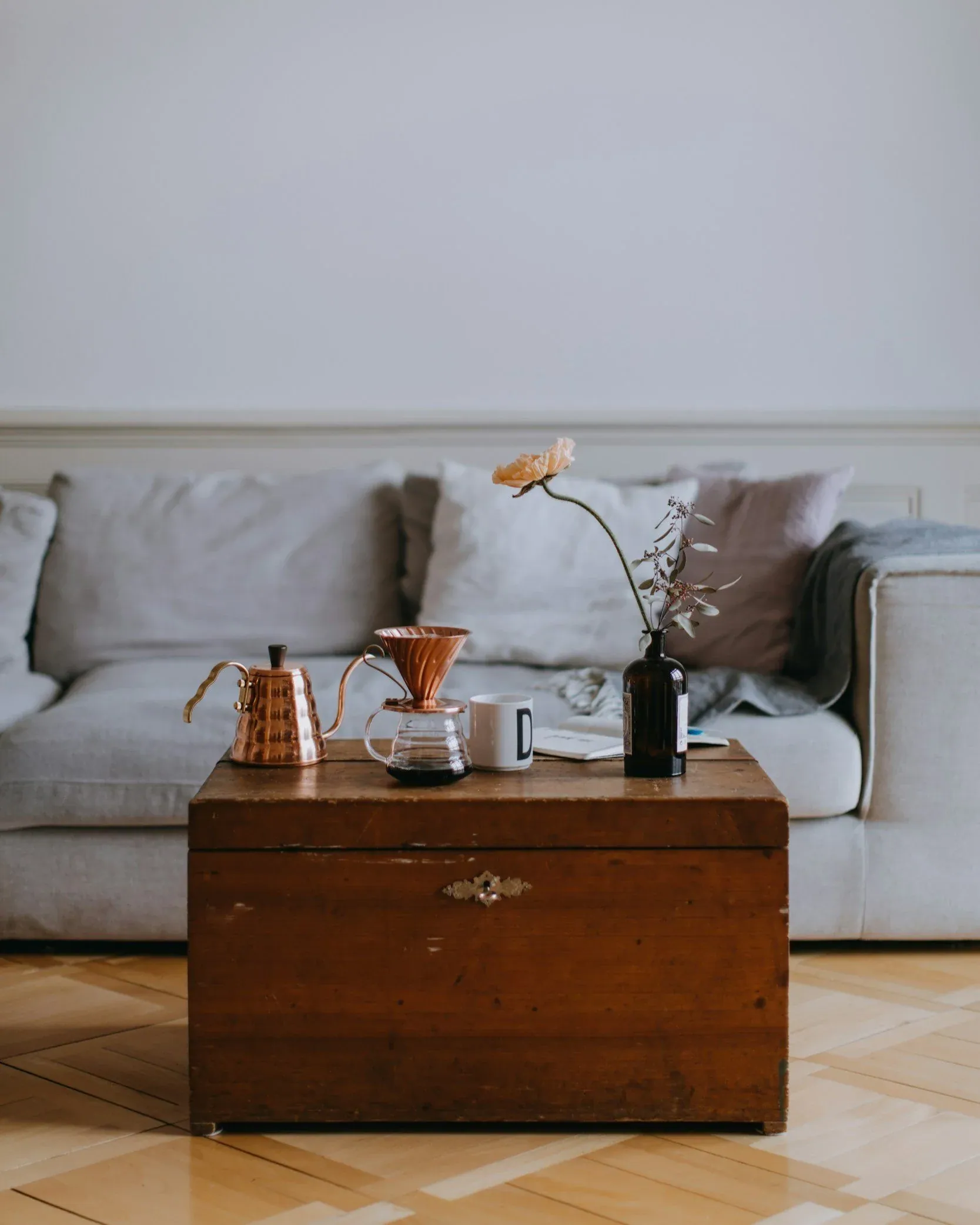 coffee dripper setup with a copper kettle and dripper on a chest in front of a grey couch. There is also a pink flower inside a glass jug sitting on the chest.